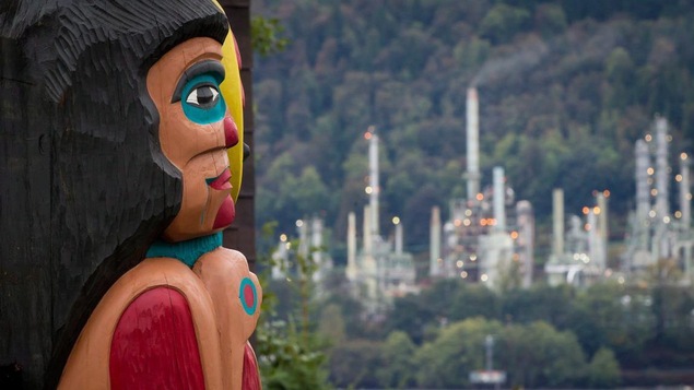 Un totem de profil avec en arrière-plan de la forêt et une usine de pétrole.