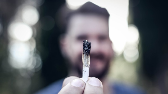 Un homme tient un joint de cannabis entre ses doigts et il le montre en avant de lui.