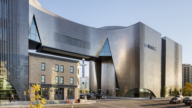Un édifice moderne en verre et briques avec passerelle construit autour d'un vieil hôtel.