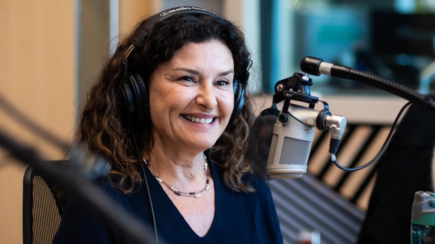 Une femme, casque d'écoute sur la tête, sourit devant le micro d'un studio de radio lors d'une entrevue. 