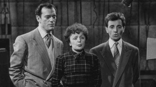 Eddie Constantine, Édith Piaf et Charles Aznavour chantent devant une caméra sur un plateau de télévision.
