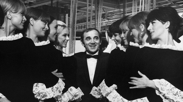 Charles Aznavour se tient debout au milieu de huit jeunes femmes à ses côtés.