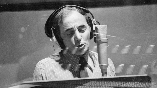 Charles Aznavour chante dans un micro, un casque sur les oreilles, dans un studio de musique.