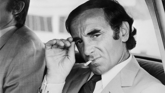 Charles Aznavour à l'arrière d'une voiture, le regard sombre qui fixe la caméra, une cigarette aux lèvres
