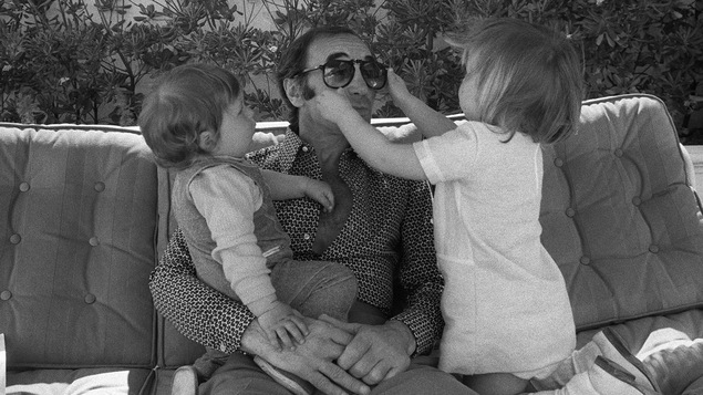 Charles Aznavour sur un canapé tient son garçon sur ses genoux et sa fille à ses côtés joue avec ses lunettes de soleil.