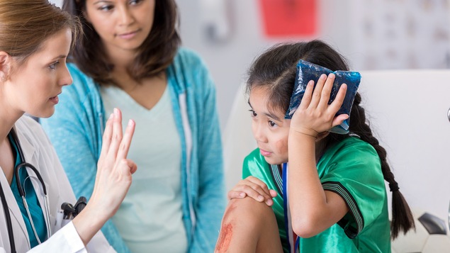 Une jeune joueuse de soccer tient un sac de glace sur sa tête, tandis qu'une médecin lui pose des questions. La mère de la jeune fille se trouve en arrière-champ.