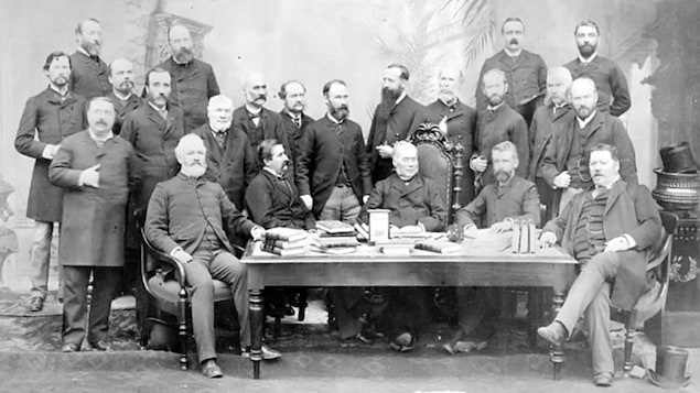 Les dirigeants de cinq provinces du Canada se rencontrent à Québec en 1887. Ils posent en veston devant une table où se trouvent des piles de livres.