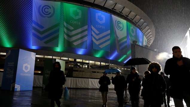 Des personnes marchent sous la pluie devant un stade orné de banderoles annonçant la COP24.