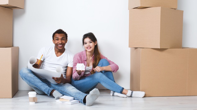 Un jeune couple mixte est assis par terre et sourit. Il est entouré de boîtes de déménagement car il arrive dans une nouvelle maison.