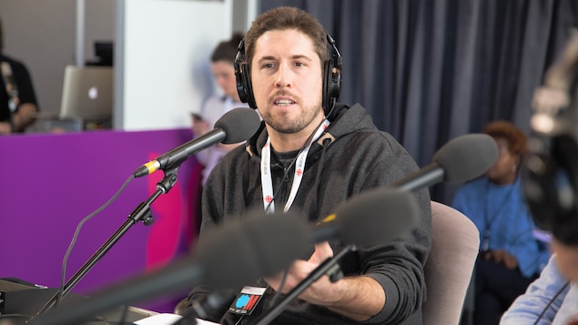 L'auteur est assis derrière un micro et lève le bras gauche en parlant.