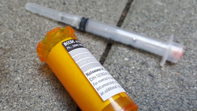 Une seringue usée et un flacon ouvert sur lequel est apposé une ordonnance pour Naloxone sont sur le pavé.