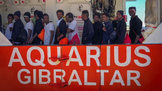 Une dizaine de migrants font la file sur le pont de l'Aquarius avec le nom du bateau peint sur le flanc du navire.