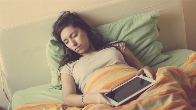 Une jeune femme dort un lit avec sa tablette électronique entre les mains.