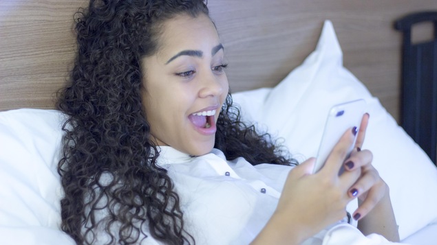 Une jeune femme observant, amusée et surprise, le contenu de son téléphone mobile. Elle est au lit, vêtue d'un chemisier blanc.