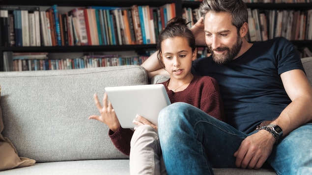 Une jeune adolescente montre quelque chose à son père sur une tablette. Les deux sont assis sur un fauteuil gris.