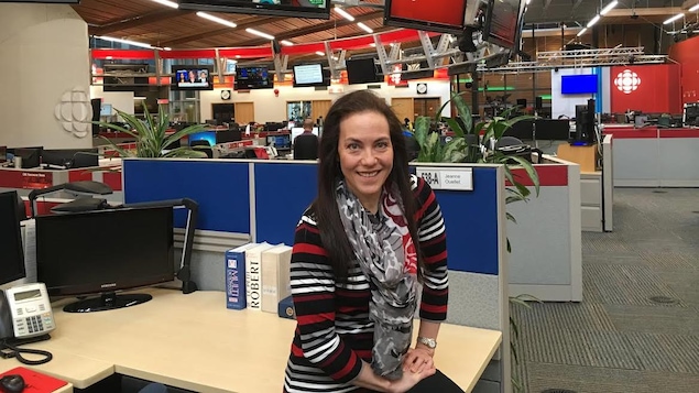 Une femme est assit sur un coin de bureau, les bras appuyés sur sa cuisse, dans une salle de nouvelles.