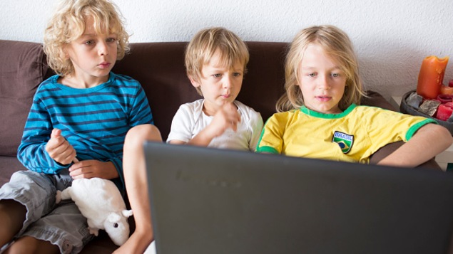 Trois jeunes enfants assis sur un divan regarde l'écran d'un ordinateur portable.