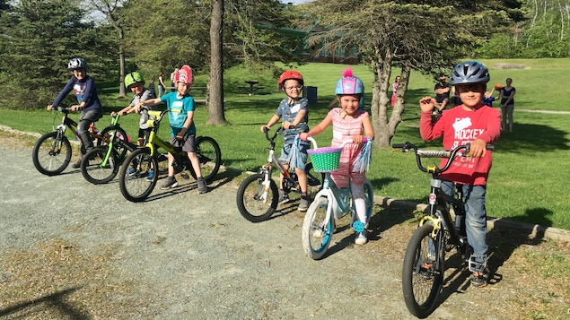 Six enfants, cinq garçons et une fille, à cheval sur leur bicyclette. Ils portent tous un casque. Ils sont dans un parc sur un sol rocheux. Ils sont arrêtés, les pieds au sol, et regardent la caméra.