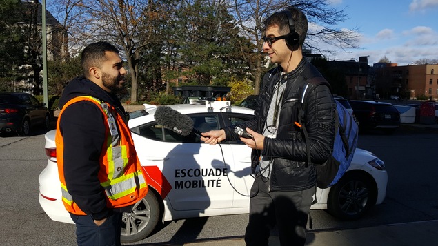 Un homme portant un dossard orange parle dans un micro tenu par un autre homme, devant une voiture sur laquelle on peut lire « Escouade mobilité ».