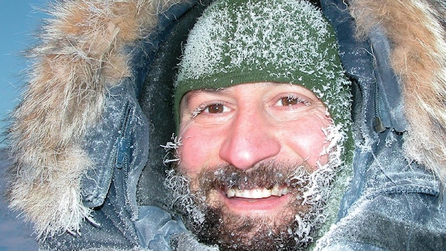 Gros plan du visage d'un homme en habits d'hiver, la barbe, les cils et la tuque recouverts de givre.