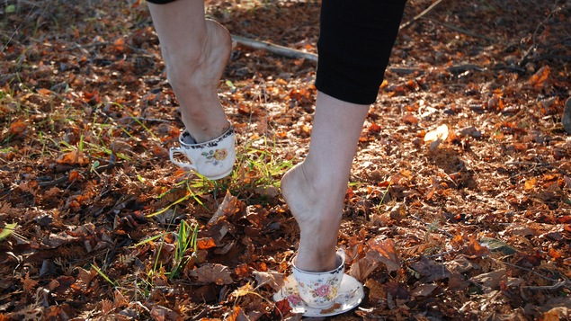 Les pieds d'une femme qui marchent dans un amas de feuilles mortes, la femme a les deux pieds dans des tasses de thé qui lui servent de souliers.
