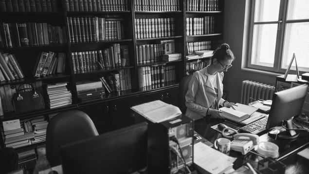 Une jeune secrétaire feuillette un livre, assise devant un écran d'ordinateur.