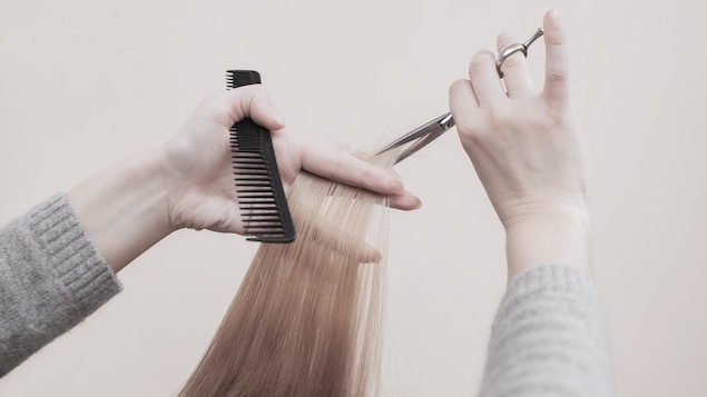 Une coiffeuse tient un peigne dans une matin, un ciseau dans l'autre et s'apprête à couper une mèche de cheveux châtains.