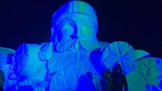 Deux personnes se font prendre en photo devant une sculpture de glace d'un homme barbu avec une tuque.
