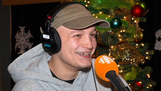 Assis derrière un micro, il porte une casquette et des écouteurs. Un sapin de Noël se profile derrière lui.