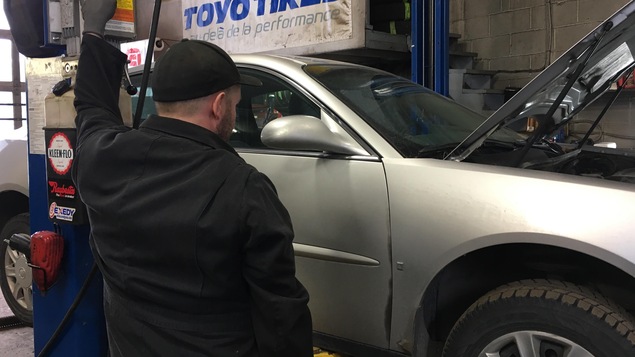 Un garagiste regarde une automobile grise qui est soulevée par un élévateur dans un garage.