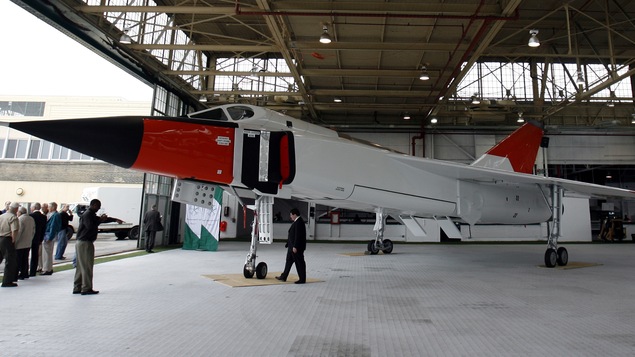 Des journalistes observent une réplique de l'avion Avro Arrow dans un hangar.