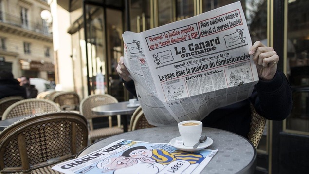 Attablé à un café français typique, un homme lit <i>Le canard enchaîné</i>, le visage caché par le journal grand ouvert entre ses mains.