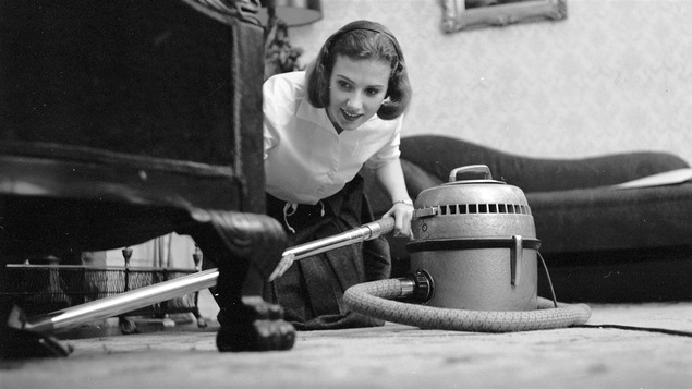 Une femme accroupie sur le sol passe l'aspirateur sous un meuble en souriant légèrement.