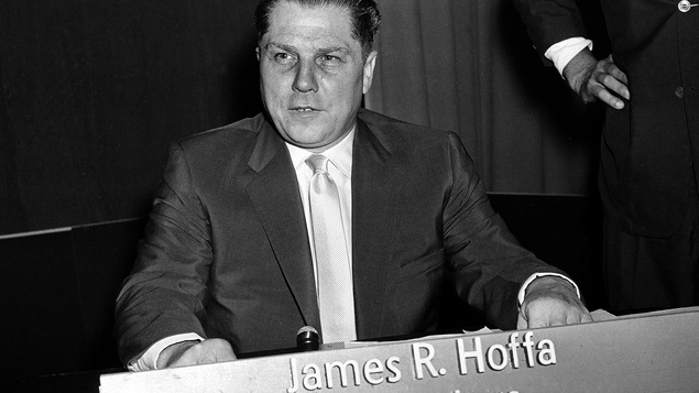 Photo en noir et blanc d'un homme assis devant une affiche avec l'inscription James R. Hoffa Teamsters.
