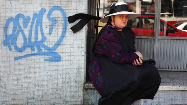 Une mennonite assise devant la vitrine d'un commerce, à côté d'un graffiti bleu illisible, regarde la caméra.