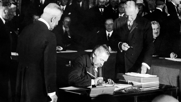 Photo en noir et blanc d'un homme moustachu qui signe un document assis. Deux hommes debout l'entourent dans une salle avec d'autres hommes.