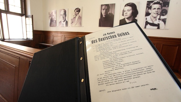 Un texte écrit en allemand est en avant-plan dans une cour de justice. Six photos sont dans le mur en arrière plan.