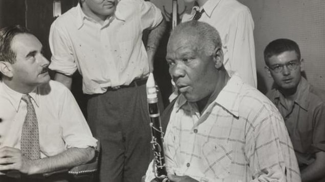 Un homme assis tient un saxophone dans ses mains. Quatre hommes, dont deux debouts, l'entourent. Un homme assis tient un saxophone dans ses mains. Quatre hommes, dont deux debouts, l'entourent.