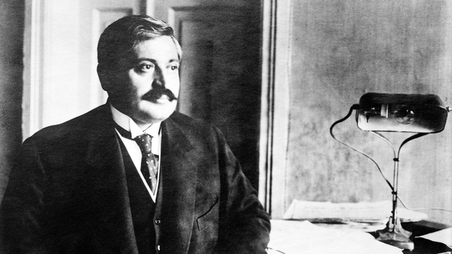 Photo en noir et blanc d'un homme moustachu qui regarde au loin et est assis à un bureau.