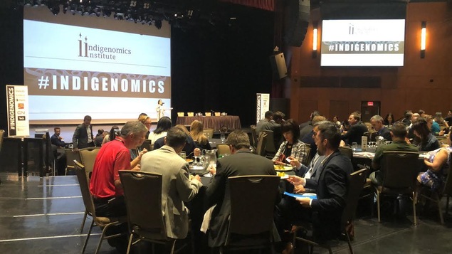 Salle où l'on voit plusieurs grandes tables rondes avec des gens qui mangent et qui assistent à une conférence du Indigenomics Institute.