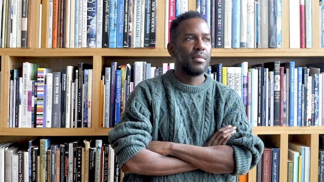 Un homme est debout les bras croisés, avec une bibliothèque derrière lui.