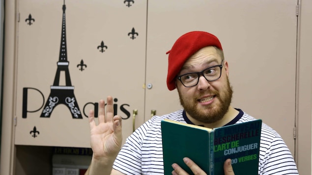Joe Vanderhelm est coiffé d'un bérét rouge et porte un t-shirt. Il tient dans sa main le livre : L'art de conjuguer. Il est devant un placard sur lequel il est écrit Paris.