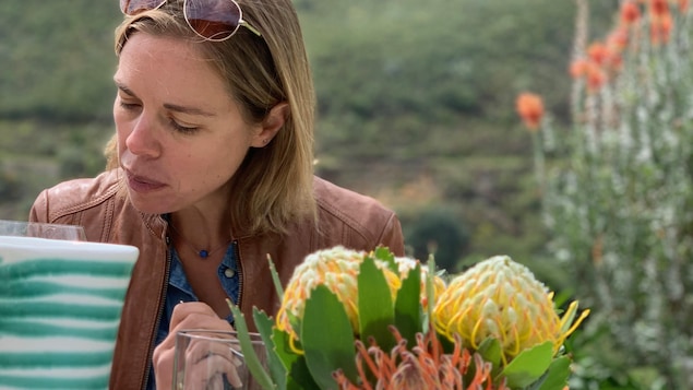 Une femme est attablée avec au premier plan un vase de fleur et en arrière-plan un paysage montagneux verdoyant.