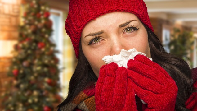 Une femme portant une tuque et des gants rouges se mouche avec un mouchoir devant un sapin.