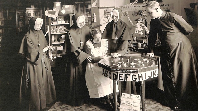 Photo en noir et blanc montrant une jeune fille entourée de trois religieuses et d'un abbé, devant un plateau affichant des lettres de l'alphabet.