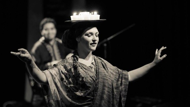 Une photo en noir et blanc montre une performance scénique de Marilyne Gélinas où elle tient un plateau de chandelles en équilibre sur sa tête