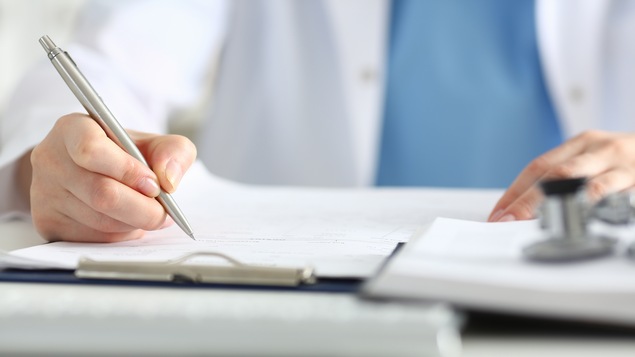 Gros plan sur le stylo tenu par un médecin en sarrau, qui remplit un formulaire.
