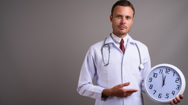 Dans un studio au mur gris, un homme médecin tient une horloge dans sa main gauche. Il la pointe de sa main droite.