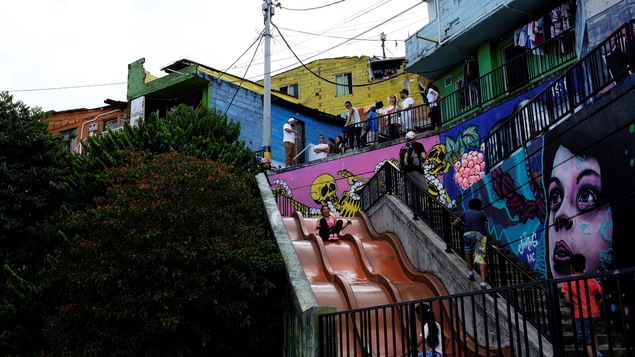 Des touristes en haut d'un enchevêtrement d'habitations dans un quartier défavorisé, ils regardent des enfants qui glissent sur des toboggans. Il y a plusieurs peintures murales très colorées.