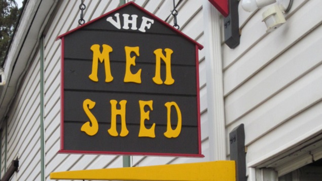 Une affiche en forme de maison sur laquelle il est écrit Men Shed en lettres jaunes.